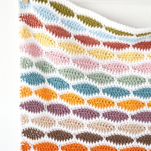 May include: A colorful, handmade crochet blanket with a repeating pattern of leaf-shaped designs. The blanket features a variety of colors, including red, orange, yellow, pink, blue, green, and brown, all set against a white background.