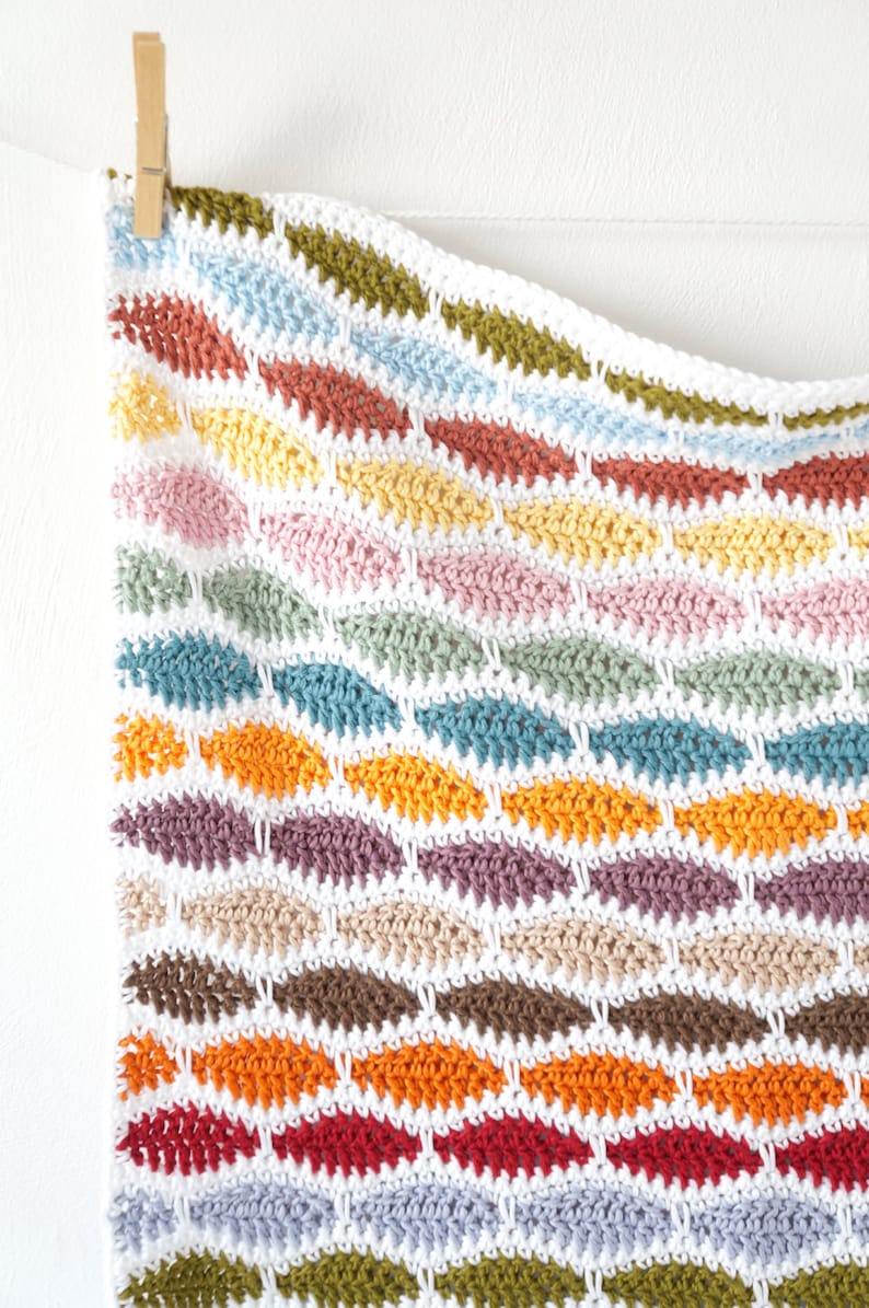 May include: A colorful, handmade crochet blanket with a repeating pattern of leaf-shaped designs. The blanket features a variety of colors, including red, orange, yellow, pink, blue, green, and brown, all set against a white background.