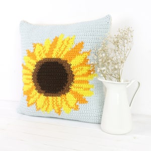 Puede incluir: Una almohada de crochet con un dise&ntilde;o de girasol. El girasol es amarillo y naranja con un centro marr&oacute;n. La almohada es azul claro.