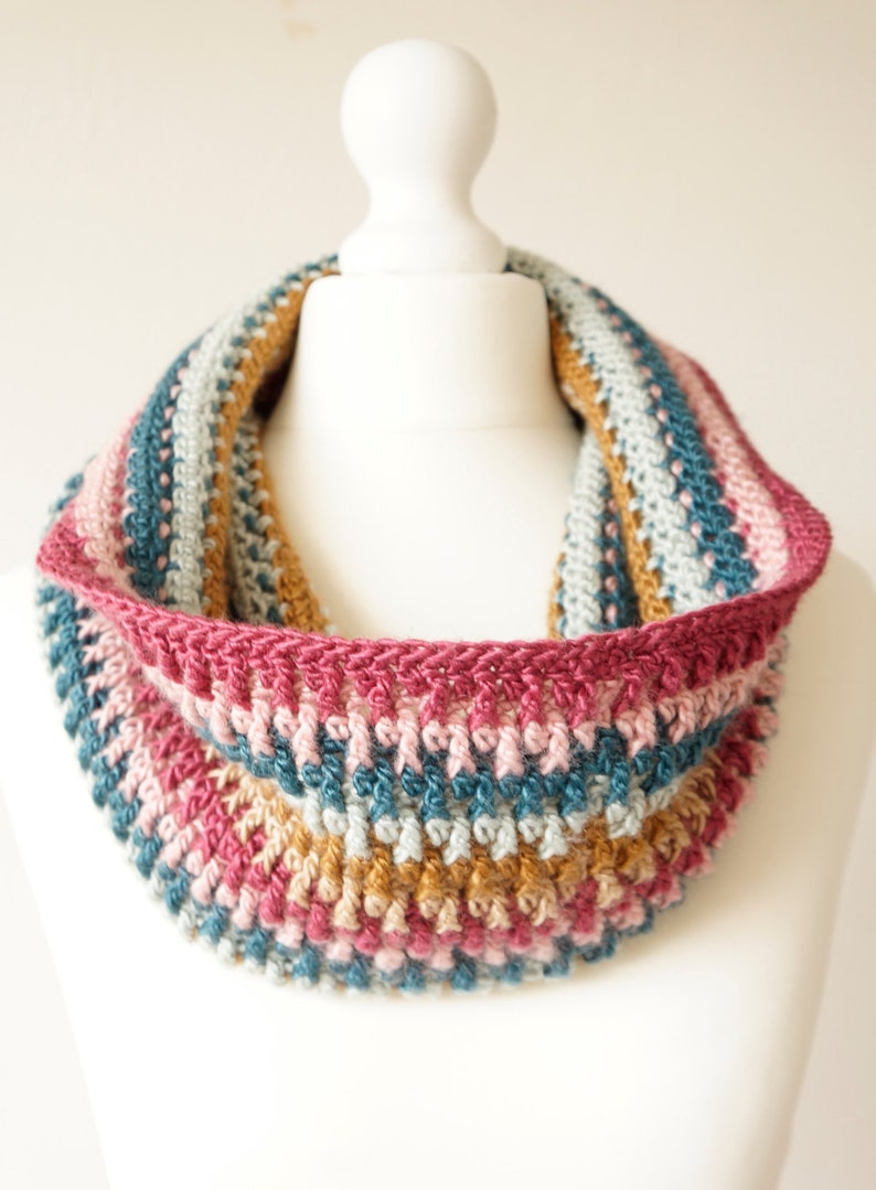 Puede incluir: Una capucha de ganchillo con un patr&oacute;n de rayas multicolores en tonos de rosa, azul y marr&oacute;n. La capucha est&aacute; hecha con un punto texturizado y tiene un dise&ntilde;o simple y elegante.
