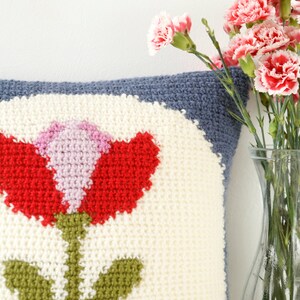 Puede incluir: Una almohada blanca de crochet con un dise&ntilde;o de flor roja, rosa y verde. La almohada tiene un borde azul y est&aacute; sobre una superficie blanca.