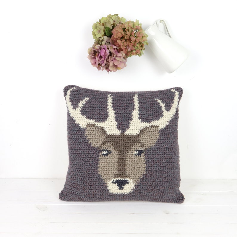 May include: A gray crocheted pillow with a brown deer face and antlers. The deer has white eyes and a white nose.