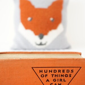 May include: An orange book with the title "Hundreds of Things a Girl Can Make" printed in black on a white triangle.