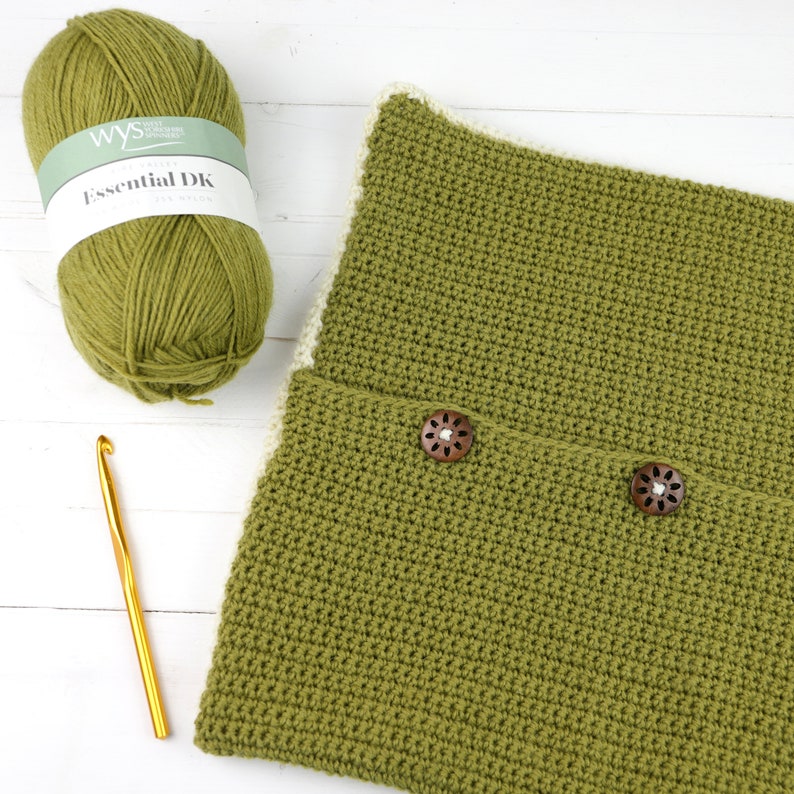 May include: A green crocheted cushion with two wooden buttons. The cushion has a white border. A ball of green yarn and a crochet hook are also in the image. The yarn label reads "Essential DK"