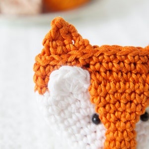 May include: Close-up of a crocheted fox toy with orange fur, white face, and black button eyes.