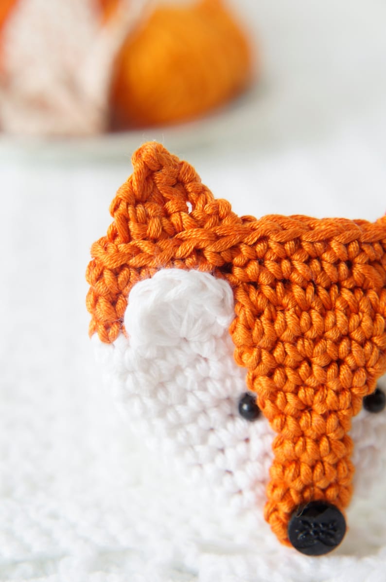 May include: Close-up of a crocheted fox toy with orange fur, white face, and black button eyes.