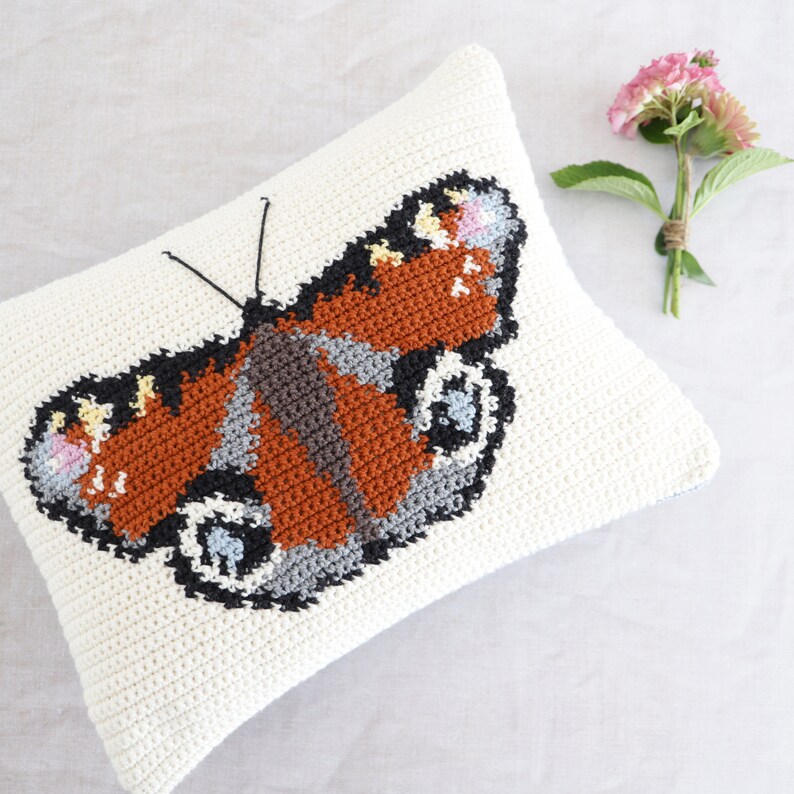 Op de afbeelding: Een wit gehaakt kussen met een kleurrijk vlinderontwerp. De vlinder heeft bruine, oranje, zwarte, grijze en blauwe accenten.