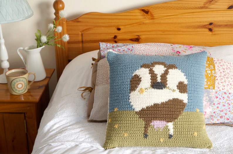 May include: A crocheted pillow with a brown and white cow design on a blue background. The cow is standing in a green field with yellow flowers.