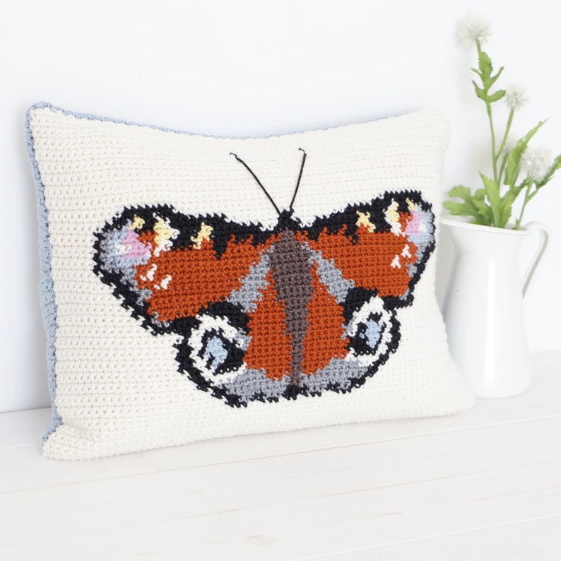 Op de afbeelding: Een wit gehaakt kussen met een kleurrijk vlinderontwerp. De vlinder heeft bruine, oranje, zwarte, grijze en blauwe accenten.