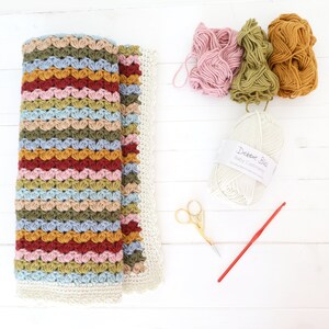May include: A crocheted baby blanket with a multi-colored striped pattern in shades of pink, blue, green, and brown. The blanket is folded in half and has a white crocheted border. There are balls of yarn in pink, green, and brown, a ball of white yarn labeled "Debbie Bliss Baby Cashmerino", a pair of gold scissors, and a red crochet hook.