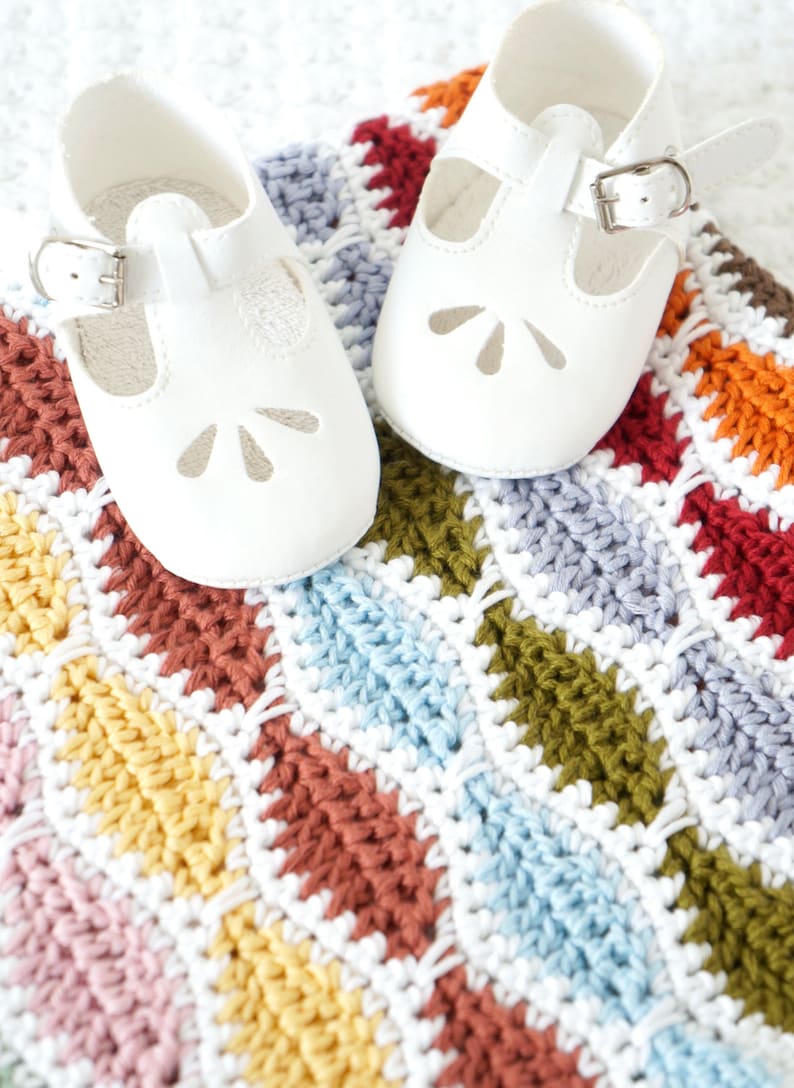 May include: A pair of white baby shoes with a T-strap design and buckle closure, resting on a colorful, hand-crocheted blanket. The blanket features a repeating pattern of various colors including red, orange, yellow, and blue.