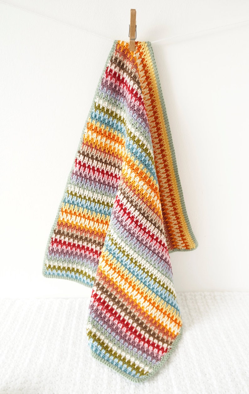 May include: A colorful striped crochet baby blanket with a textured stitch pattern. The blanket is hanging from a wooden clothespin.