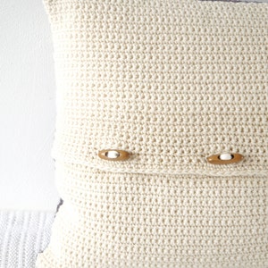 May include: A white crocheted pillow with two wooden buttons. The pillow is on a white surface.