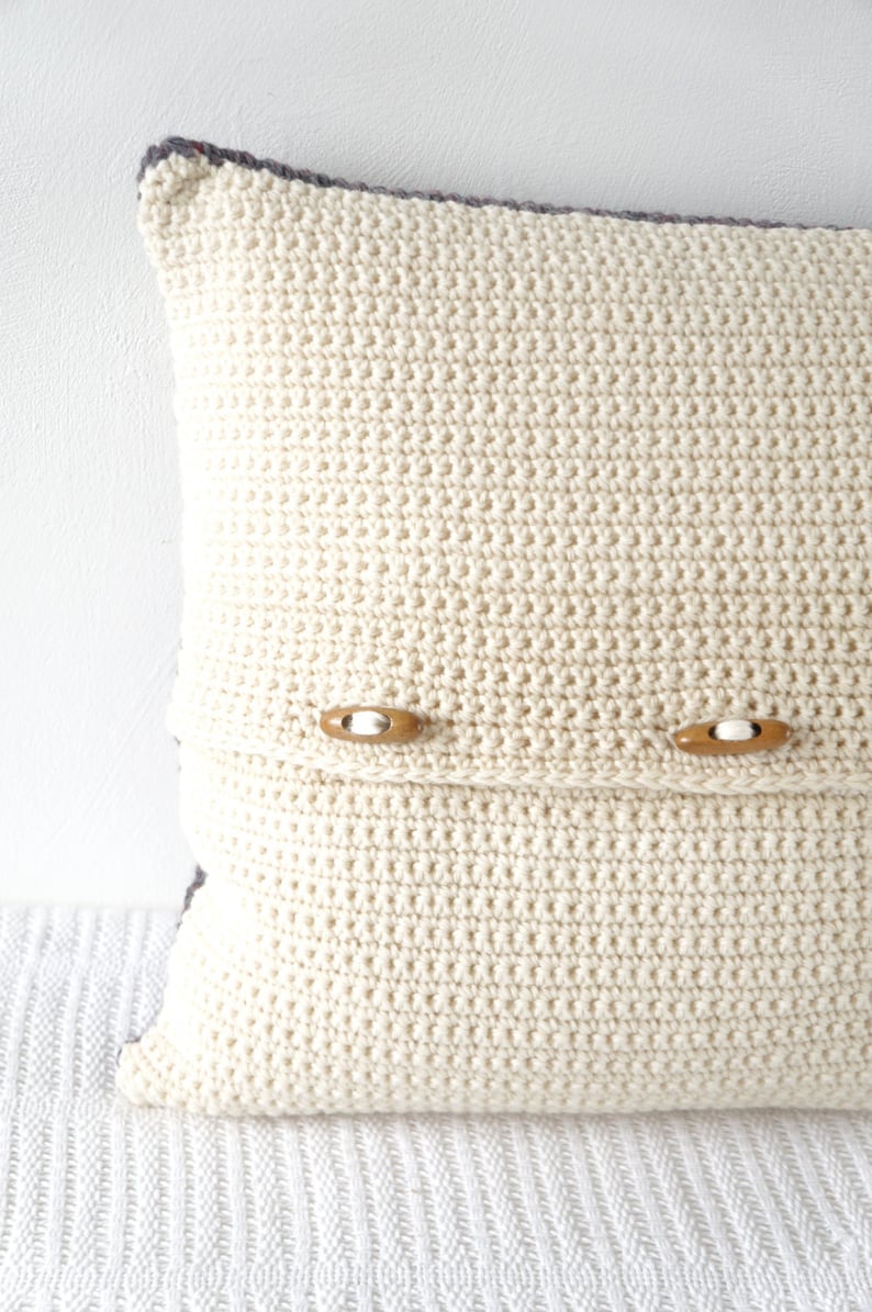 May include: A white crocheted pillow with two wooden buttons. The pillow is on a white surface.