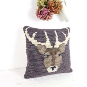 May include: A gray crocheted pillow with a brown deer face and white antlers. The pillow is on a white surface.