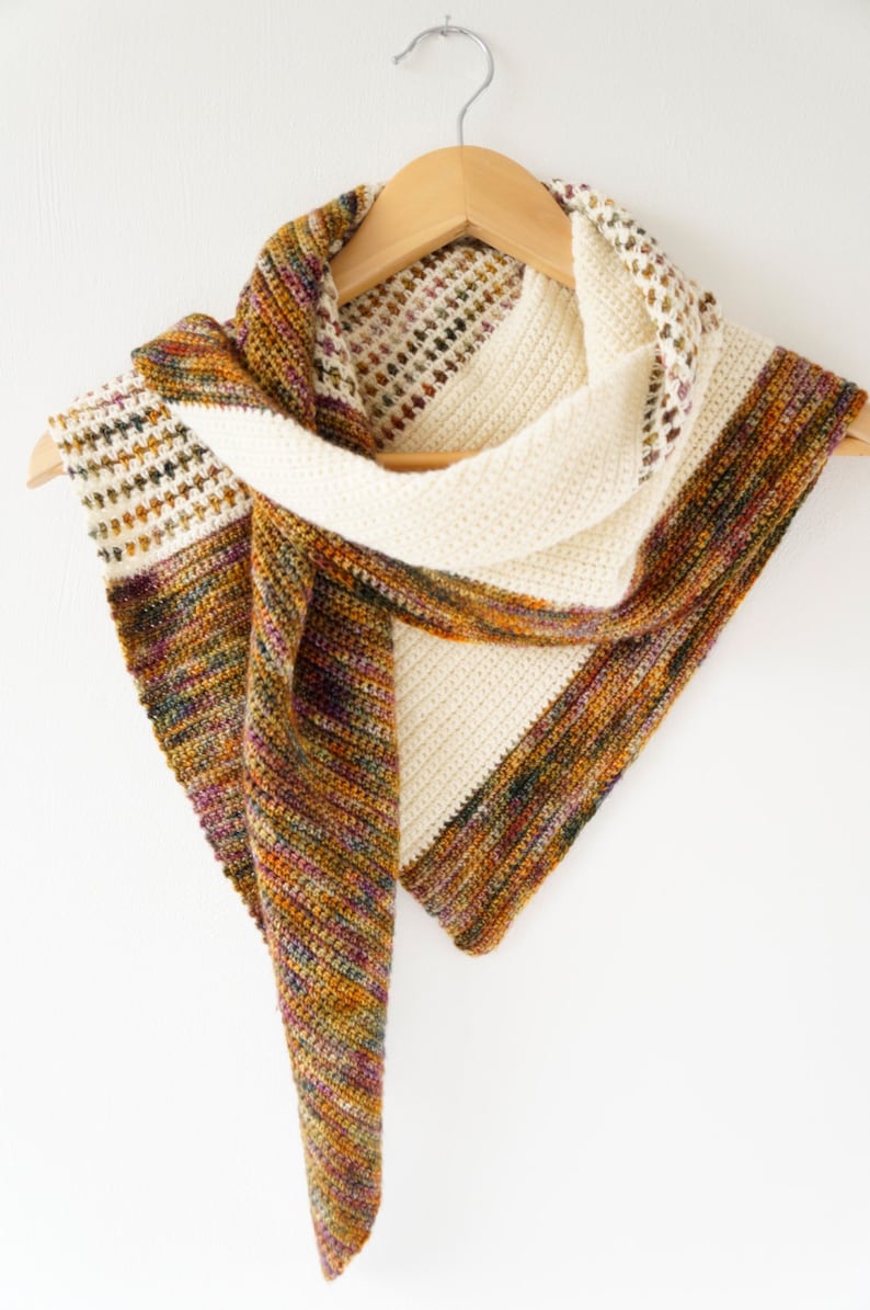 May include: A crocheted shawl with a white and multi-coloured design. The shawl is hanging on a wooden hanger.
