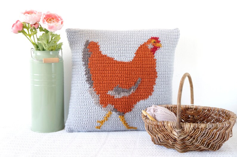 May include: A crocheted pillow with a gray background and an orange chicken design. The pillow is sitting on a white surface next to a wicker basket and a vase of pink flowers.