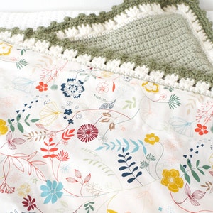 Puede incluir: Una manta de beb&eacute; de crochet verde con un borde de crochet blanco. La manta tiene un estampado floral sobre un fondo blanco.
