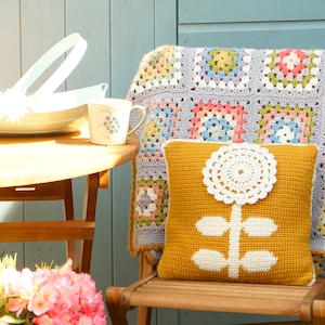 May include: A crocheted granny square throw blanket in shades of pink, blue, and green is draped over a wooden chair. A yellow crocheted pillow with a white flower design sits on the chair.