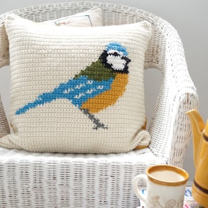 May include: A white crocheted pillow with a bluebird design. The bird has blue, green, yellow, and white accents. The pillow is sitting on a white wicker chair.