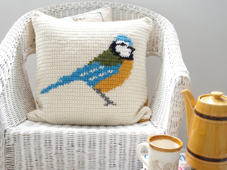 May include: A white crocheted pillow with a bluebird design. The bird has blue, green, yellow, and white accents. The pillow is sitting on a white wicker chair.