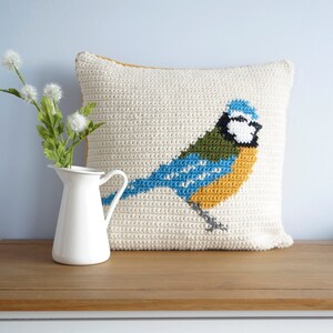 May include: A crocheted pillow with a bluebird design. The bird is yellow, blue, green, and white. The pillow is cream colored and has a brown border.