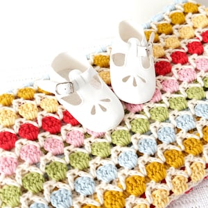 Puede incluir: Una manta de bebé de crochet blanco con un patrón colorido y multicolor. La manta está hecha con un punto de granny square y presenta una variedad de colores, incluyendo rojo, amarillo, azul, verde y rosa. Dos zapatos de bebé blancos están colocados encima de la manta.
