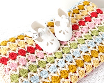 Może przedstawiać: Biały, szydełkowy kocyk dla dziecka z kolorowym, wielokolorowym wzorem. Kocyk wykonany jest ściegiem granny square i zawiera różne kolory, w tym czerwony, żółty, niebieski, zielony i różowy. Na kocyku leżą dwa białe buciki dla dziecka.