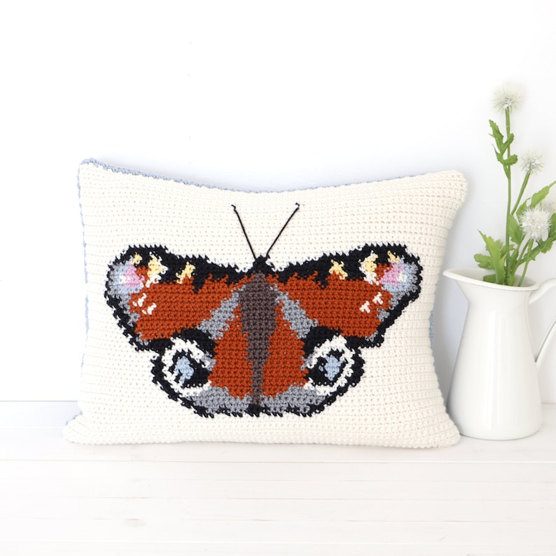 Op de afbeelding: Een wit gehaakt kussen met een bruin, zwart en blauw vlinderontwerp. De vlinder heeft een rood en oranje lichaam en zwarte en witte vlekken op zijn vleugels.