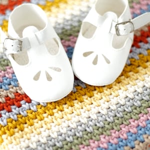 May include: A pair of white baby shoes with a strap and buckle on a colorful crocheted blanket.
