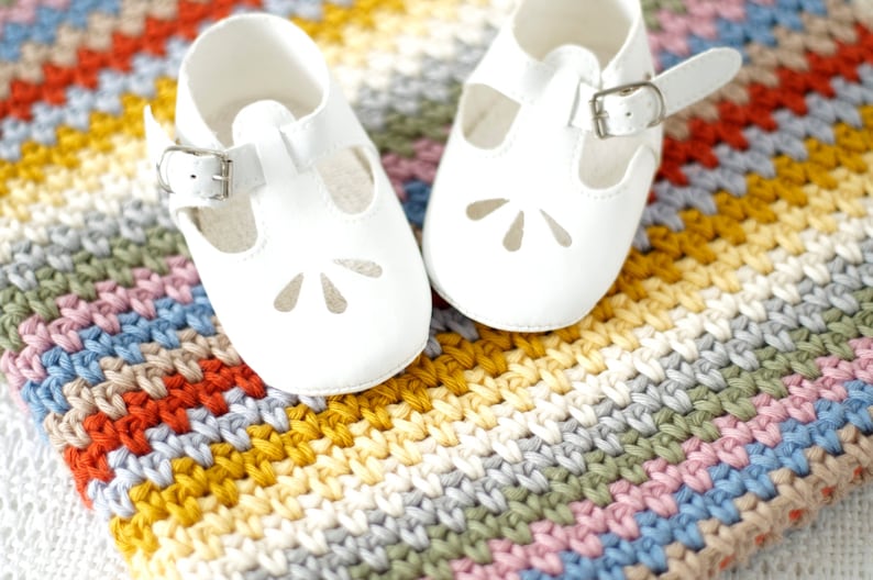 May include: A pair of white baby shoes with a strap and buckle on a colorful crocheted blanket.