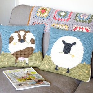 May include: Two crocheted throw pillows with animal designs. The pillow on the left features a brown and white cow with pink udders. The pillow on the right features a white sheep with a black face and a yellow flower in its mouth. Both pillows have a blue background and green grass at the bottom.