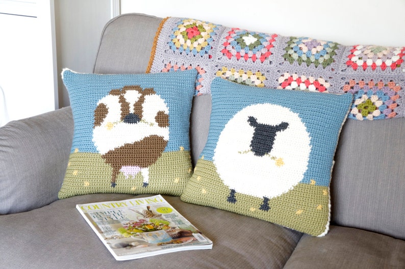 May include: Two crocheted throw pillows with animal designs. The pillow on the left features a brown and white cow with pink udders. The pillow on the right features a white sheep with a black face and a yellow flower in its mouth. Both pillows have a blue background and green grass at the bottom.
