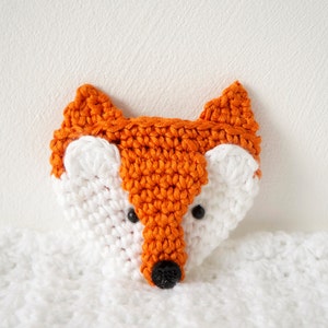 May include: A crocheted fox head with orange ears and a white face. The fox has black eyes and a black nose.
