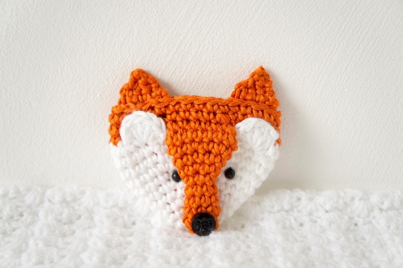 May include: A crocheted fox head with orange ears and a white face. The fox has black eyes and a black nose.