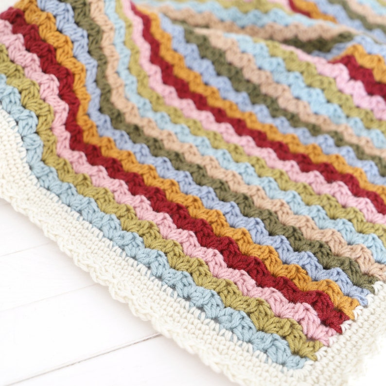 May include: A multi-colored crocheted blanket with a white border. The blanket is made with a variety of colors, including red, orange, yellow, green, blue, and purple. The blanket is made with a simple stitch pattern and has a soft, textured feel.