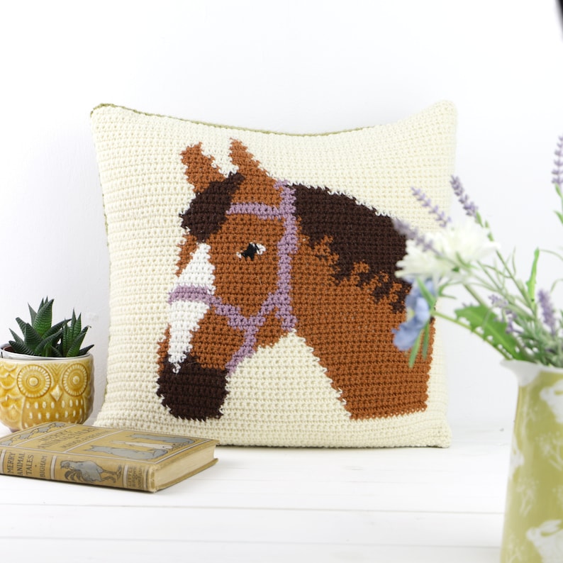 May include: A crocheted cushion featuring a brown horse head design. The horse is wearing a purple bridle. The cushion is on a white surface with a book and a vase of flowers.