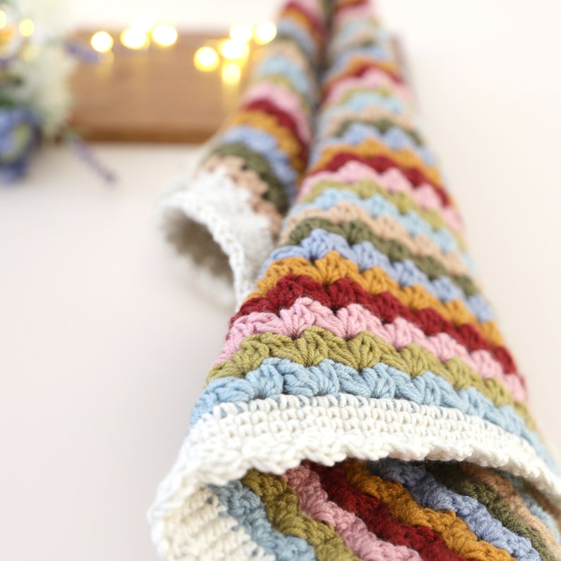 May include: A colorful crocheted baby blanket with a white border. The blanket is made with a variety of colors, including pink, blue, green, and yellow.