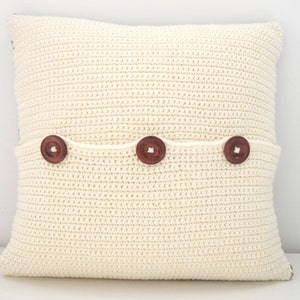 May include: A white crocheted pillow with three brown buttons. The pillow has a pocket on the back.