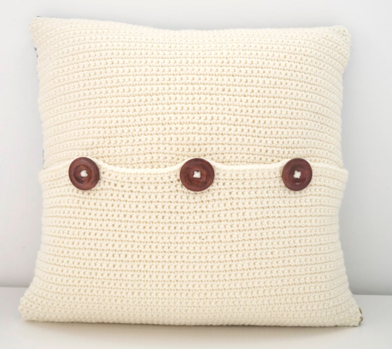 May include: A white crocheted pillow with three brown buttons. The pillow has a pocket on the back.