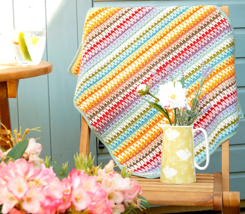 May include: A colorful crocheted throw blanket with a diagonal pattern in shades of red, orange, yellow, green, blue, and purple. The blanket is draped over a wooden chair with a yellow pitcher and white flowers.