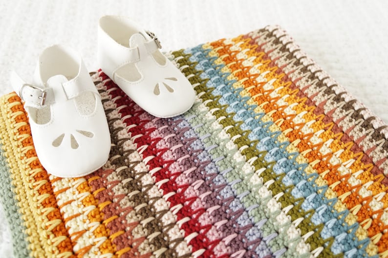 May include: A colorful striped crocheted baby blanket with two white baby shoes on top.