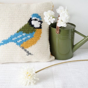 May include: A white crocheted pillow with a bluebird design. The bird is yellow, blue, and green. The pillow is sitting on a white surface with a green watering can and white flowers.