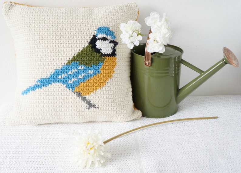 May include: A white crocheted pillow with a bluebird design. The bird is yellow, blue, and green. The pillow is sitting on a white surface with a green watering can and white flowers.