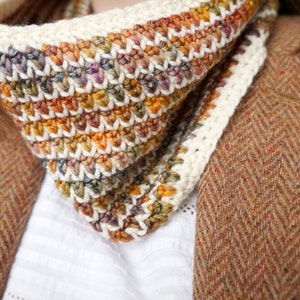 May include: A close-up of a crocheted neck warmer with a white border and a multi-colored pattern of orange, brown, and green. The neck warmer is worn over a white shirt and a brown tweed jacket.