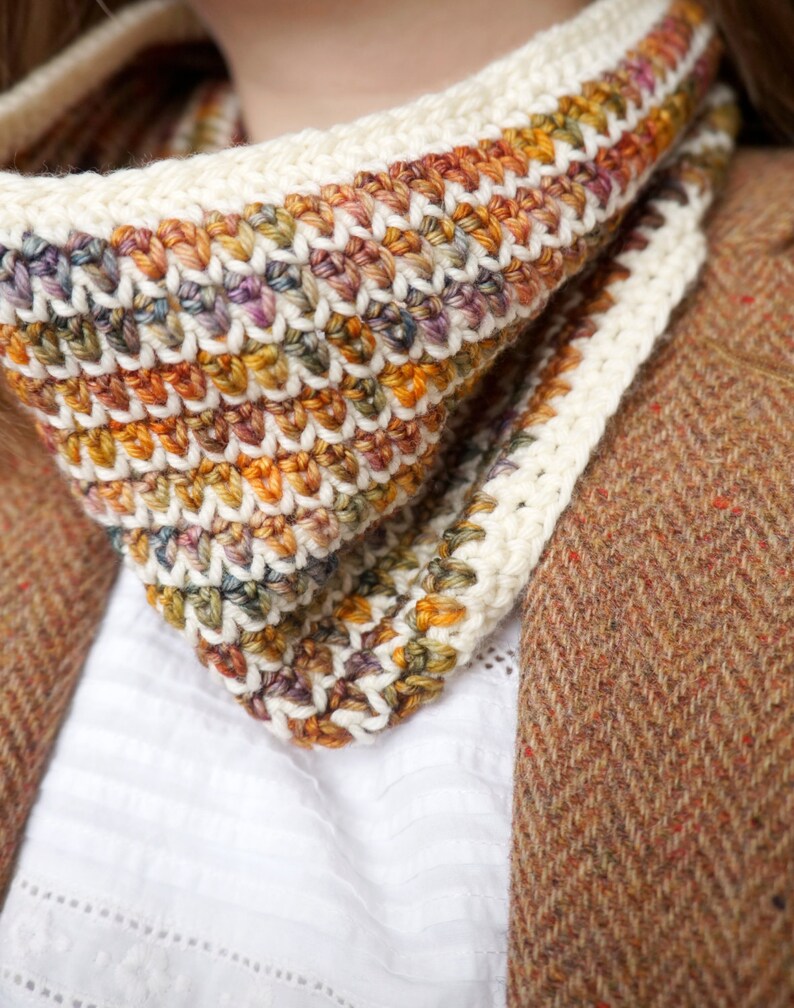 May include: A close-up of a crocheted neck warmer with a white border and a multi-colored pattern of orange, brown, and green. The neck warmer is worn over a white shirt and a brown tweed jacket.