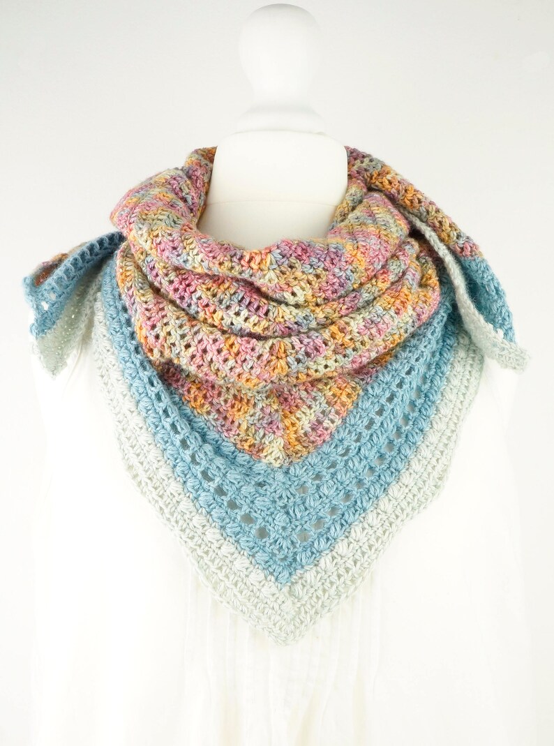 May include: A colorful, handmade crochet shawl displayed on a mannequin. The shawl features a mix of pastel colors, including pink, yellow, blue, and gray. The triangular design is visible, with a textured stitch pattern.