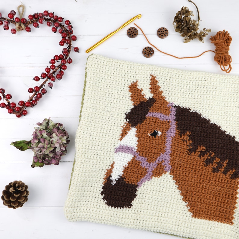 May include: A crocheted cushion featuring a brown horse head design. The horse has a purple bridle and a white nose. The cushion is on a white wooden surface.