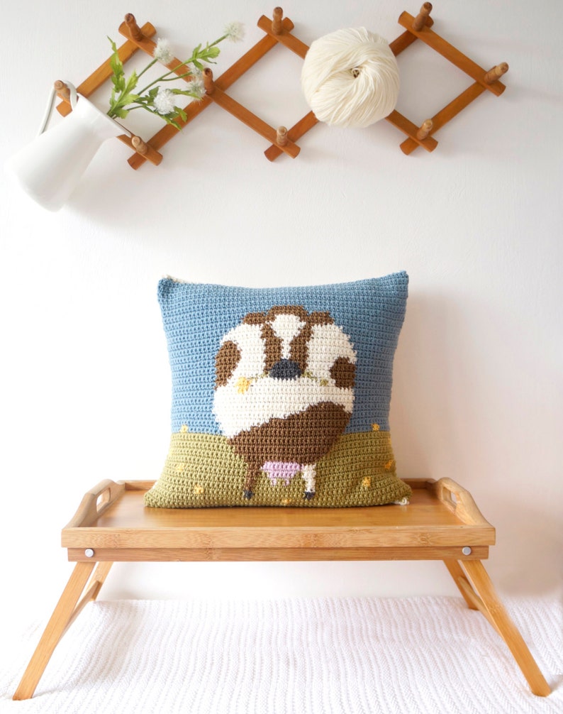 May include: A crocheted pillow with a brown and white cow design on a blue background. The cow is standing in a green field with yellow flowers. The pillow is sitting on a wooden tray.