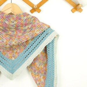 May include: A colorful, handmade crochet shawl in pastel shades of pink, yellow, and blue. The shawl is triangular, with a light blue border and a decorative openwork design. It hangs on a wooden hanger against a white background.